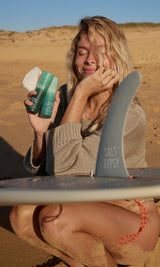 Woman holding SAMI skincare cleansers and reusable rounds beside a surfboard on the beach for cleansing outdoors